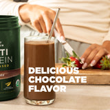 Glass of chocolate smoothie with a protein powder container and strawberries on a table.