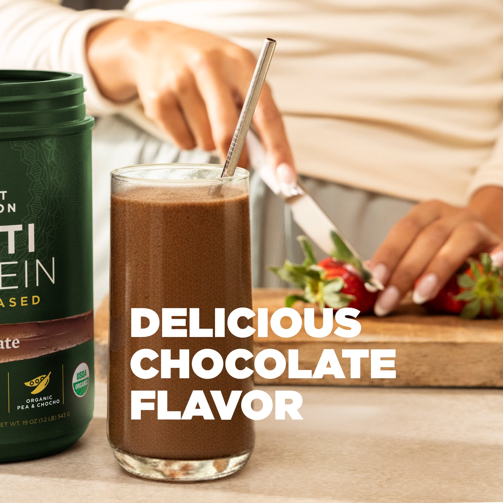 Glass of chocolate smoothie with a protein powder container and strawberries on a table.