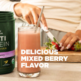 Glass of mixed berry smoothie with a protein powder container, strawberries, and a person in the background.