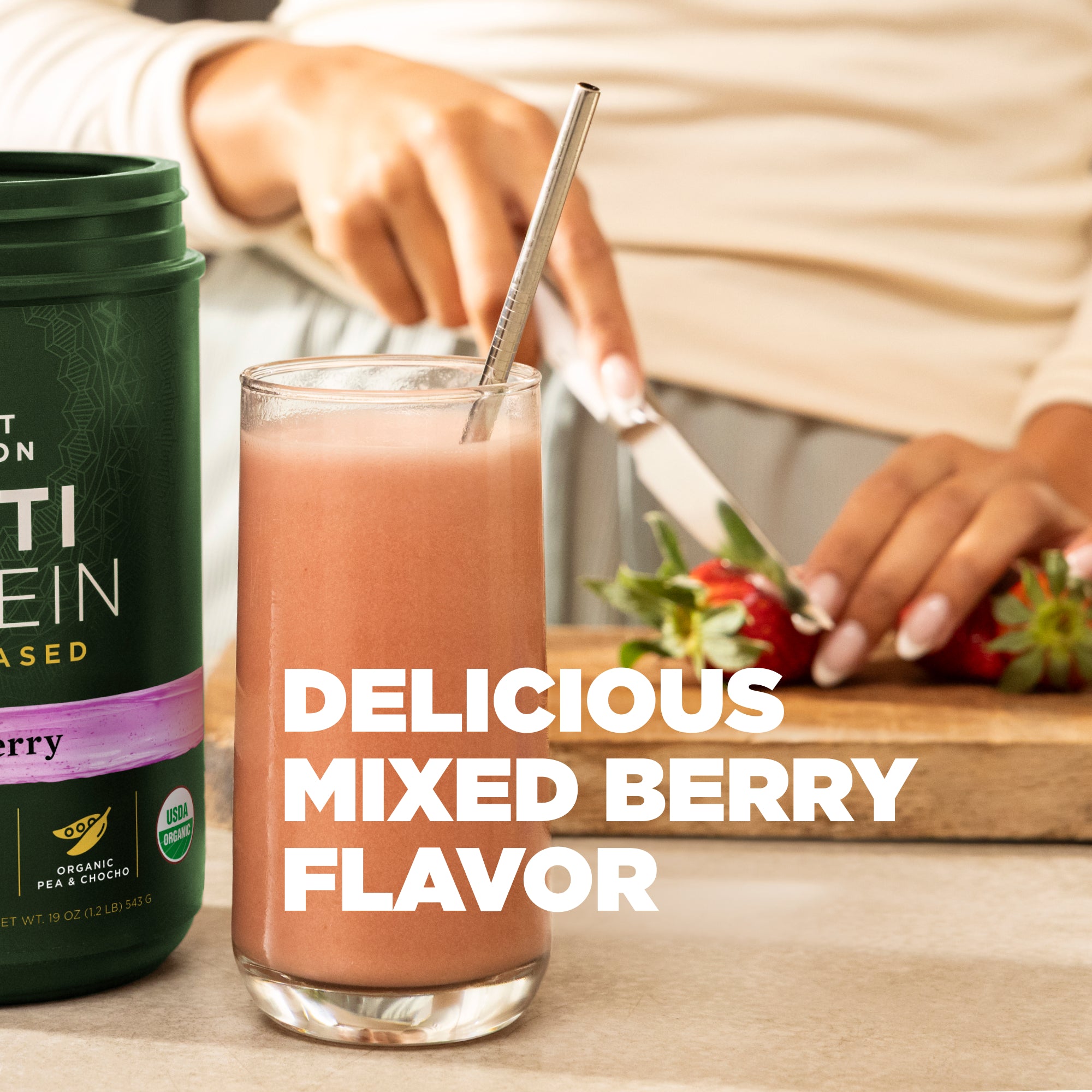 Glass of mixed berry smoothie with a protein powder container, strawberries, and a person in the background.