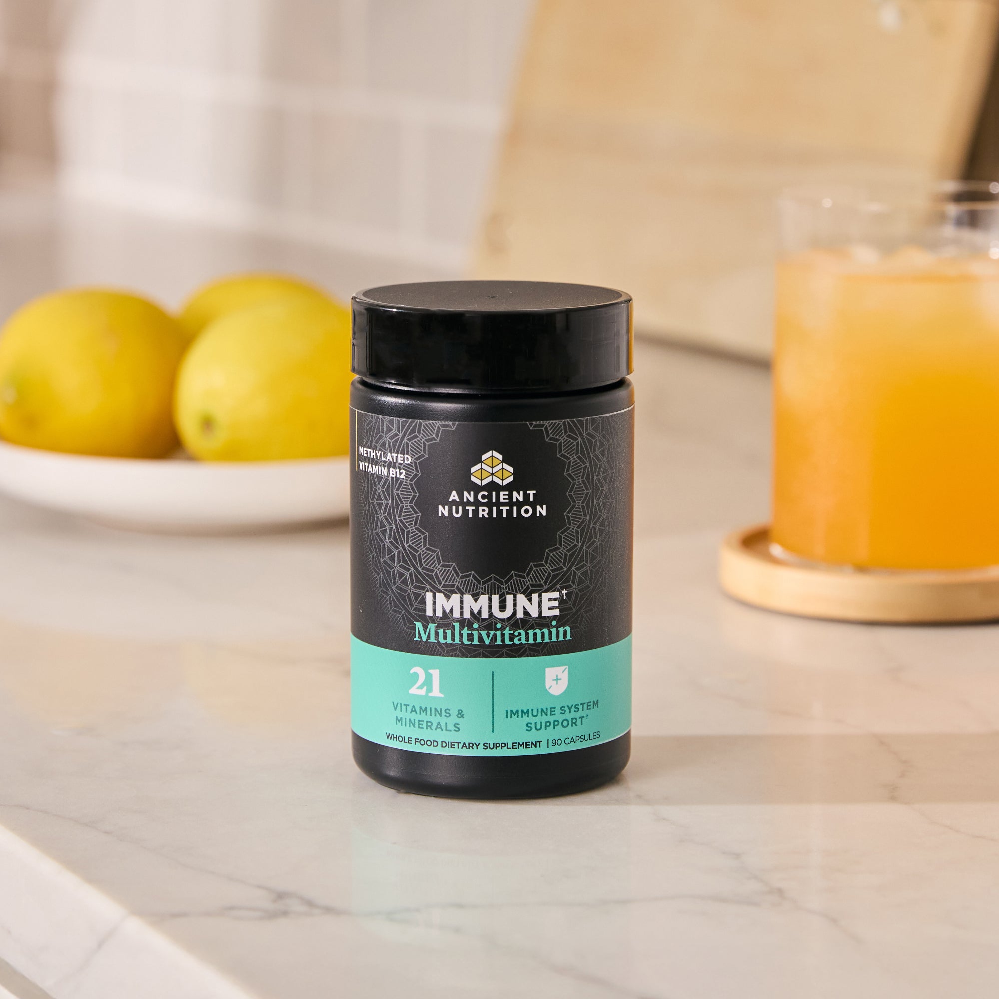 Black supplement jar labeled 'Immune Multivitamin' by Ancient Nutrition on a kitchen counter with lemons and juice.