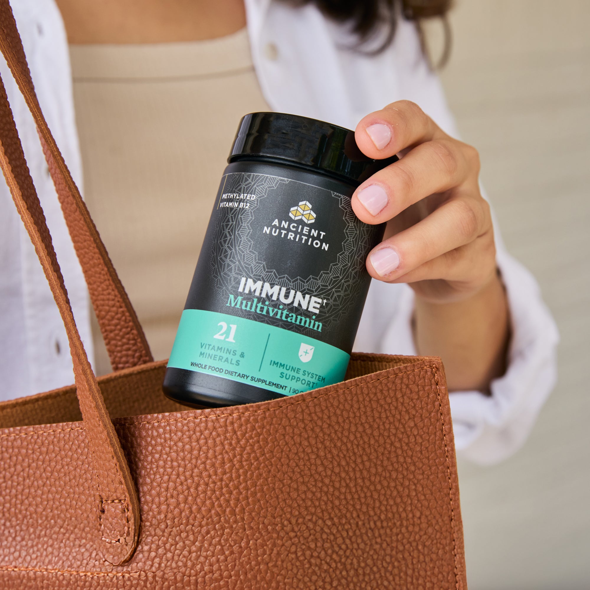Person holding a supplement container labeled 'Immune Multivitamin' by Ancient Nutrition in a brown leather bag.