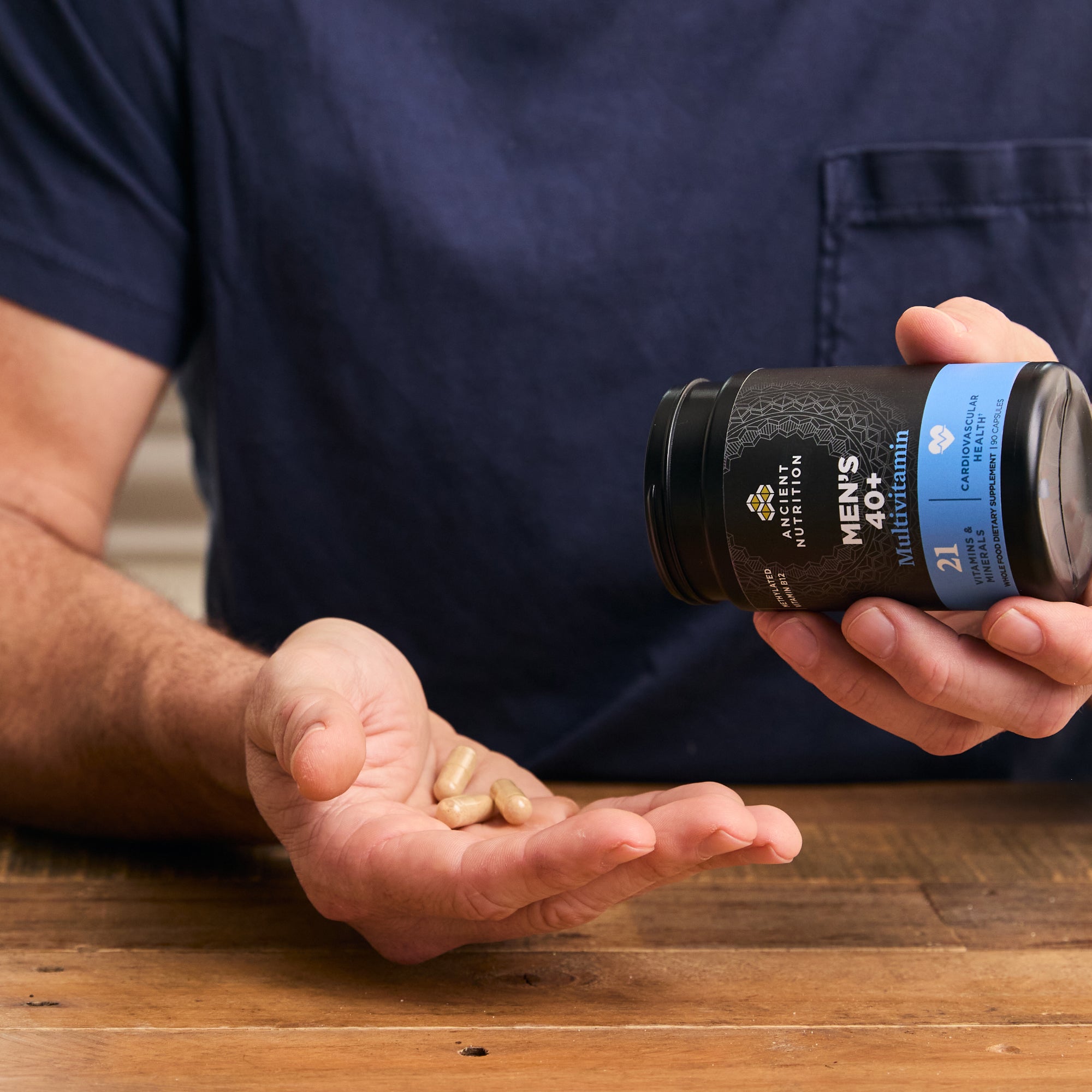 Person holding a supplement bottle labeled 'Men's Nutrition' with pills in their hand on a wooden surface.