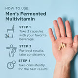 Hand holding capsules with instructions on how to use Men's Fermented Multivitamin on a light blue background