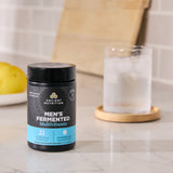 Ancient Nutrition Men's Fermented Multivitamin container on a kitchen counter with a glass of water and lemon.