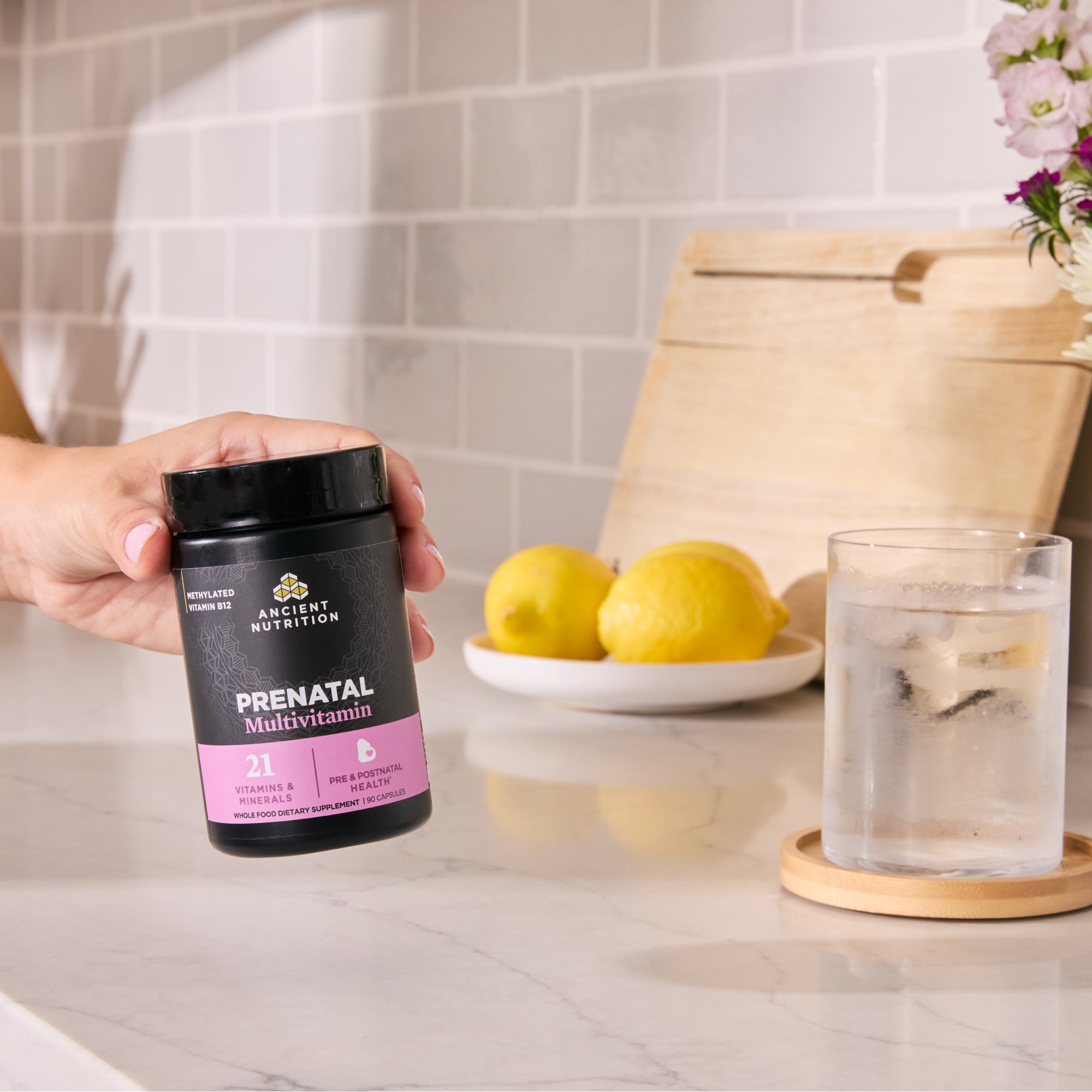 Person holding a prenatal multivitamin container on a kitchen counter with lemons and water.