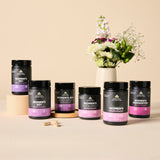 Six black supplement bottles from Ancient Nutrition with purple and pink labels are arranged on a beige surface with a floral arrangement in the background. The bottles include Women's 50+, Women's 40+, Women's 40+ once daily, Women's once daily, Women's multivitamin, and Women's Fermented.