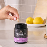 Hand holding a supplement capsule above a 'Women's 40+' multivitamin container on a kitchen counter.