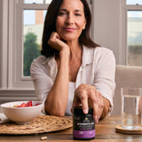 Woman holding a supplement bottle on a table with a bowl of fruit and glass of water.