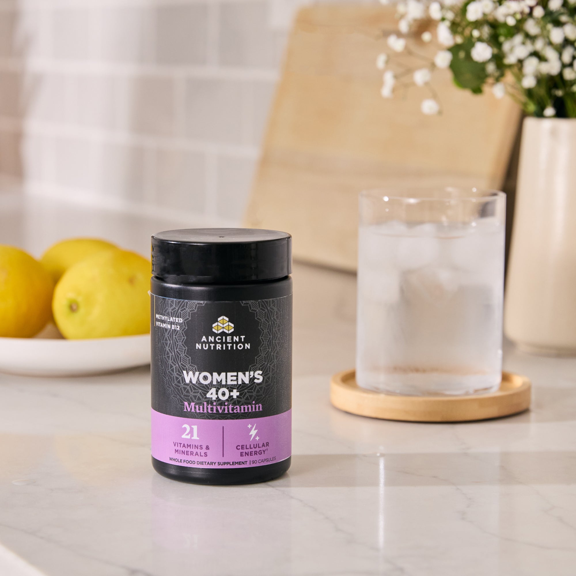 Ancient Nutrition Women's 40+ Multivitamin bottle on a kitchen counter with lemons and a glass of water in the background.