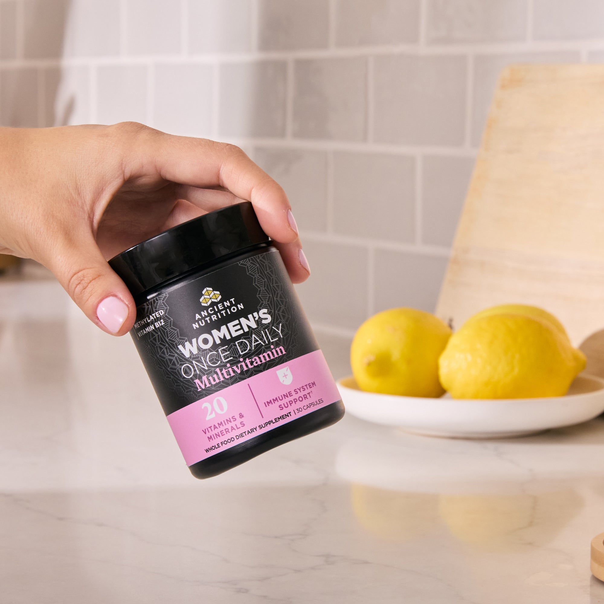 Hand holding a supplement container labeled 'Women's Once Daily Multivitamin' with lemons in the background.