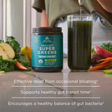 Container of Organic Super Greens with a glass of green juice and vegetables on a table.