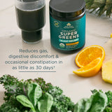 Supplement container labeled 'Super Greens' with a glass of dark liquid, oranges, and kale on a light surface.