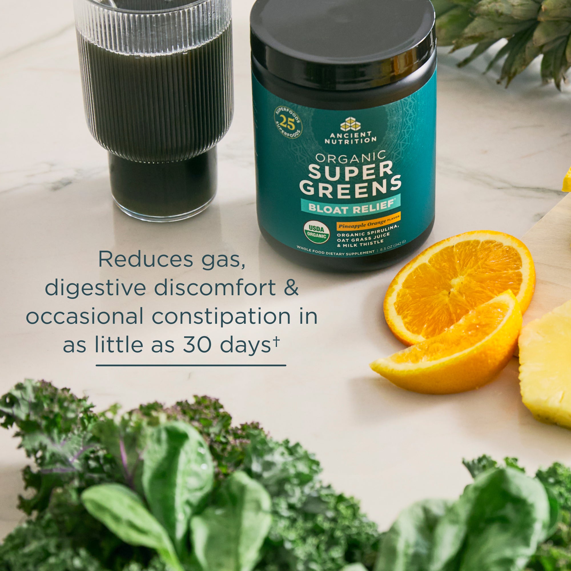 Supplement container labeled 'Super Greens' with a glass of dark liquid, oranges, and kale on a light surface.