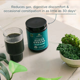 Organic Super Greens supplement jar with a glass of green juice and kale on a light surface.
