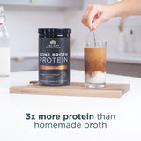 Person stirring a glass of pumpkin spice bone broth protein with a container of Ancient Nutrition Bone Broth Protein on a wooden cutting board.