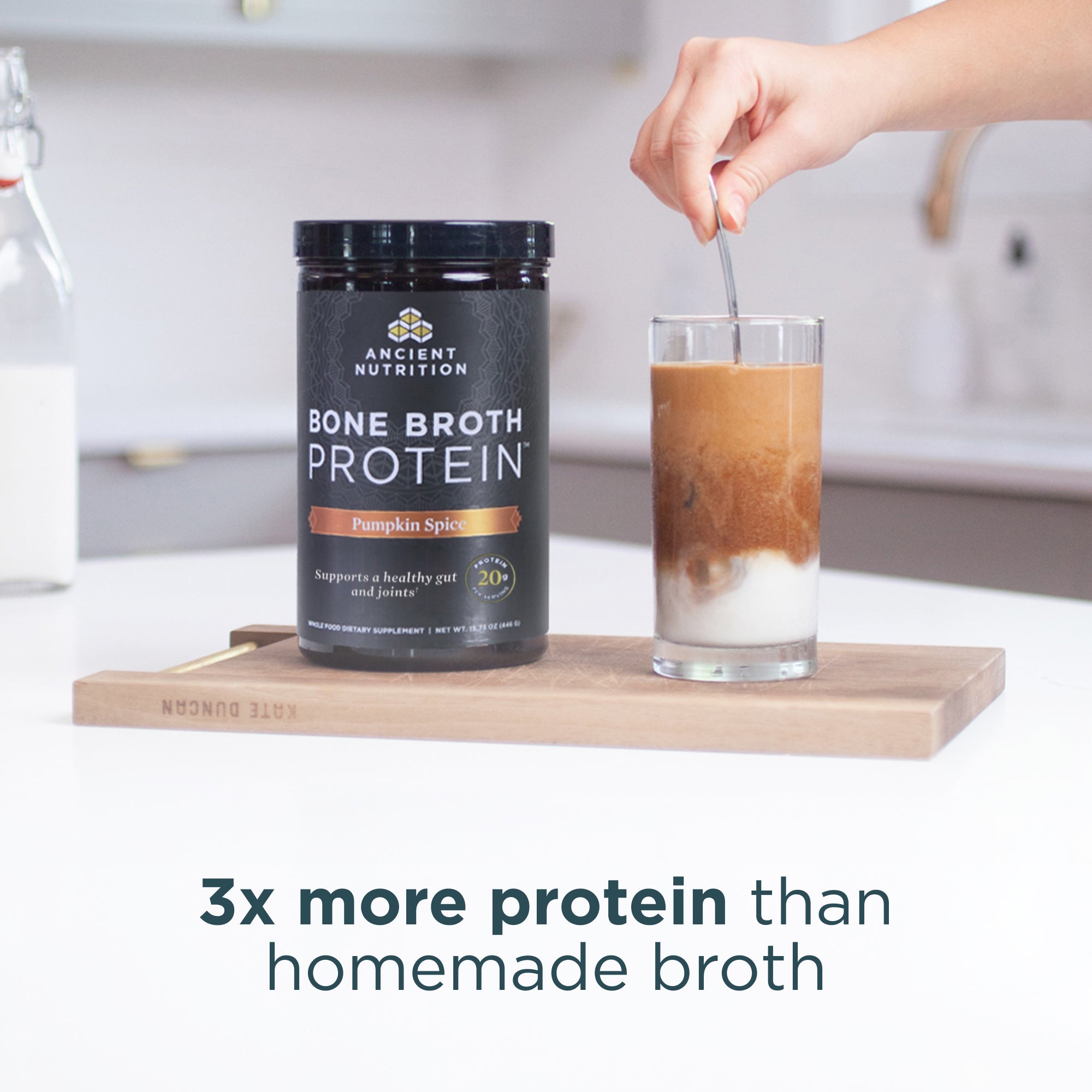 Person stirring a glass of pumpkin spice bone broth protein with a container of Ancient Nutrition Bone Broth Protein on a wooden cutting board.