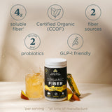 Ancient Nutrition Organic Fiber supplement tub in Pineapple Mango flavor, shown on a beige studio background with feature callouts: 4g soluble fiber, certified organic, 2 fiber sources, 2 billion probiotics, and GLP-1 friendly. A glass of yellow drink and pineapple slices are placed around the tub.