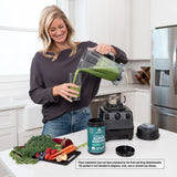 Woman pouring a green smoothie from a blender in a kitchen with Super Greens powder container.