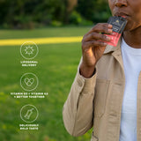 A person outdoors in a grassy park holding a small black and pink pouch of Liposomal Vitamin D3 + K2 Gel pack Pineapple . The image features three white circular icons with text overlays on the left: 'LIPOSOMAL DELIVERY', 'VITAMIN D3 + VITAMIN K2 = BETTER TOGETHER', and 'DELICIOUSLY BOLD TASTE'.