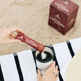 person adding multi collagen protein stick pack into coffee on the beach